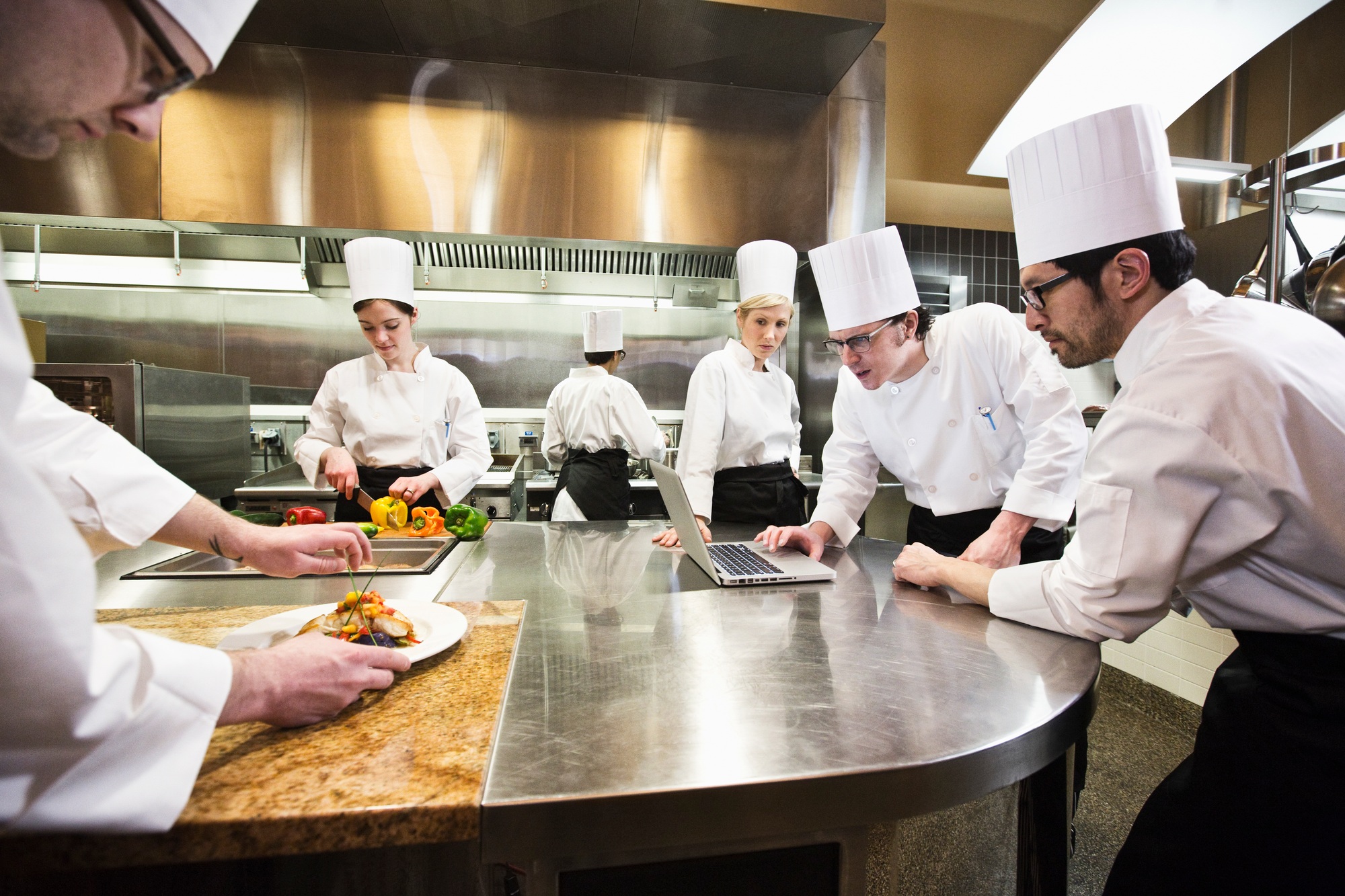 A crew of chef's working in a commercial kitchen, while several chefs wok on a laptop computer.